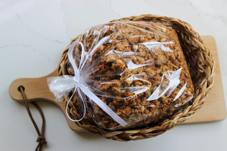 May include: A clear plastic bag of granola, tied with a white ribbon, is presented in a woven basket. The basket sits on a wooden chopping board with a handle. The granola is a mix of golden brown and tan ingredients.