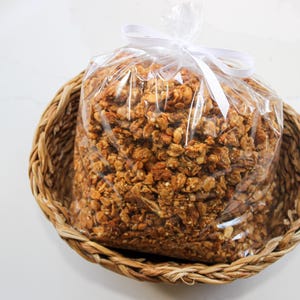 May include: A clear plastic bag filled with granola, tied with a white ribbon, sits inside a woven basket. The granola is a mix of golden brown and tan colours. The basket is made of woven, light brown material.