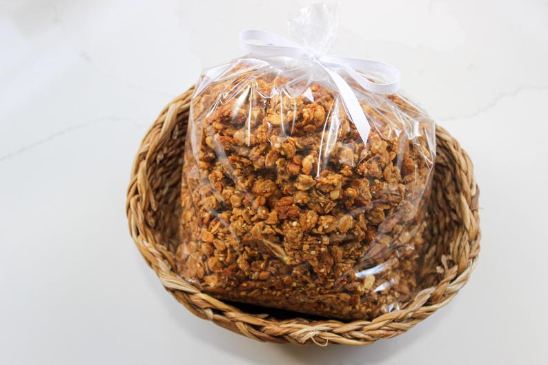 May include: A clear plastic bag filled with granola, tied with a white ribbon, sits inside a woven basket. The granola is a mix of golden brown and tan colours. The basket is made of woven, light brown material.
