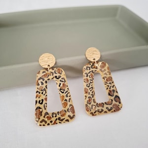 Könnte beinhalten: Ein Paar goldfarbene Ohrringe mit Leopardenmuster. Die Ohrringe haben eine rechteckige Form mit einem Ausschnitt in der Mitte und einem kleinen, strukturierten Kreis oben.