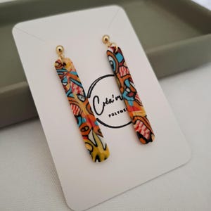Peut inclure: Une paire de boucles d'oreilles pendantes colorées. Les boucles d'oreilles sont longues et rectangulaires, avec un motif vibrant orange, jaune, bleu et rouge. Elles ont des tiges dorées. Les boucles d'oreilles sont présentées sur une carte blanche avec un logo.