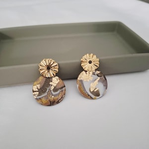 Può includere: Un paio di orecchini color oro con un design circolare. Gli orecchini presentano una parte superiore a forma di fiore color oro e un fondo circolare marmorizzato con accenti di foglia d'oro. Gli orecchini sono esposti su un vassoio grigio.