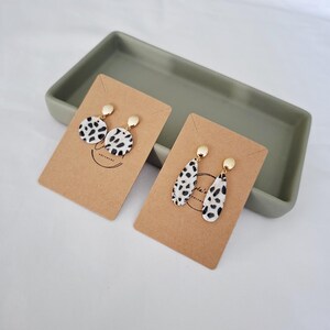Puede incluir: Dos pares de pendientes en tarjetas marrones. Uno es redondo, el otro en forma de lágrima. Ambos tienen una base blanca con manchas negras y tachuelas doradas. Los pendientes se presentan en una bandeja rectangular gris.
