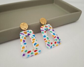 May include: A pair of earrings with a gold-colored circular stud and a white, rectangular drop. The drop features a colorful confetti pattern of red, orange, yellow, green, blue, and purple dots. The earrings are displayed on a gray tray.