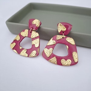 Può includere: Un paio di orecchini color magenta con dettagli dorati a forma di cuore. Gli orecchini hanno un design geometrico triangolare con un ritaglio centrale. Gli orecchini sono su una superficie bianca.