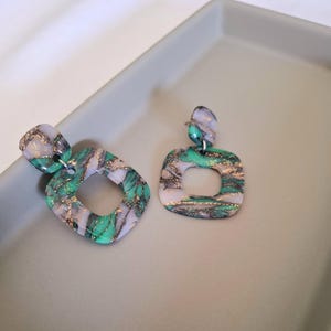 May include: A pair of square-shaped dangle earrings with a green, white, and gold marbled design. The earrings have a square opening in the center.