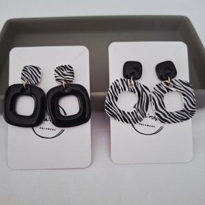Puede incluir: Dos pares de pendientes. Uno presenta un cuadrado negro con estampado de cebra en la parte superior, y el otro un cuadrado negro con estampado de cebra en el centro. Ambos pares se muestran en tarjetas blancas.