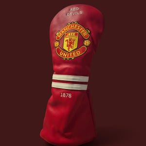 May include: Red golf club head cover with the Manchester United logo, the words "Red Devils" and "1878", and two white stripes. The cover is made of red leather.