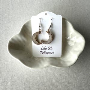 May include: A pair of crescent moon earrings with a silver-tone hook. The earrings have a light-colored, textured surface with a gold-tone trim. The earrings are displayed on a white card that reads "Lily B's Treasures".