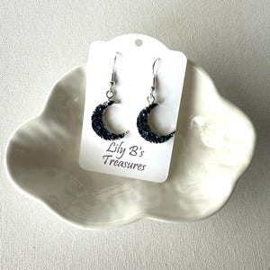 May include: A pair of crescent moon earrings with dark blue glitter, displayed on a white card that reads "Lily B's Treasures". The earrings are suspended from silver-colored hooks. The card rests on a white, cloud-shaped dish.