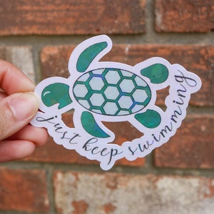 Può includere: Un adesivo bianco con un'illustrazione di tartaruga verde e blu iridescente e il testo "just keep swimming".