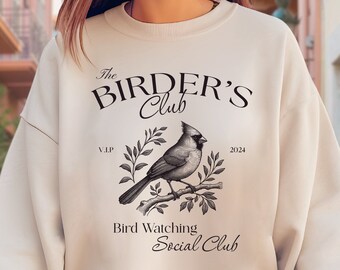 May include: A beige sweatshirt with a graphic design of a cardinal bird on a branch. The design includes the text "The Birders Club", "V.I.P", "2024", "Bird Watching", and "Social Club".