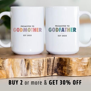 Puede incluir: Dos tazas de cerámica blanca con el texto "PROMOTED TO GODMOTHER EST 2025" y "PROMOTED TO GODFATHER EST 2025" en letras coloridas. Las tazas están sobre una superficie de madera.