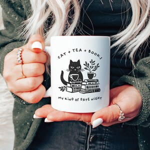 Puede incluir: Taza de cer&aacute;mica blanca con una ilustraci&oacute;n de un gato negro sosteniendo una taza de t&eacute; y sentado sobre una pila de libros. La taza tiene el texto "Cat + Tea + Books. My kind of date night."