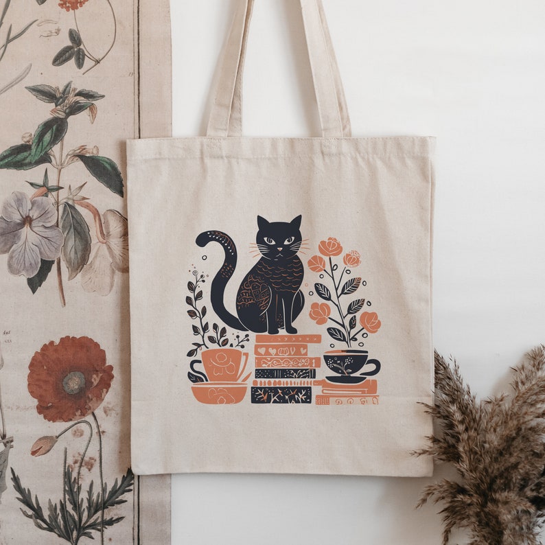 Puede incluir: Una bolsa de tela beige con un gato negro, flores naranjas y una pila de libros.