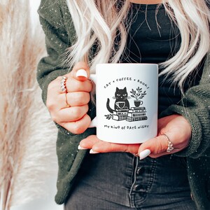 Puede incluir: Taza de cer&aacute;mica blanca con una ilustraci&oacute;n de un gato negro, una taza de caf&eacute; y una pila de libros. El texto en la taza dice "Cat + Coffee + Books. My kind of date night."
