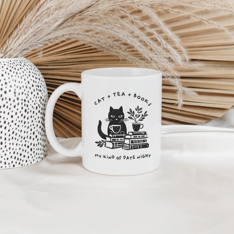 Puede incluir: Taza de cer&aacute;mica blanca con una ilustraci&oacute;n de un gato negro sentado en una pila de libros y sosteniendo una taza de t&eacute;. El texto en la taza dice "Cat + Tea + Books My Kind of Date Night".
