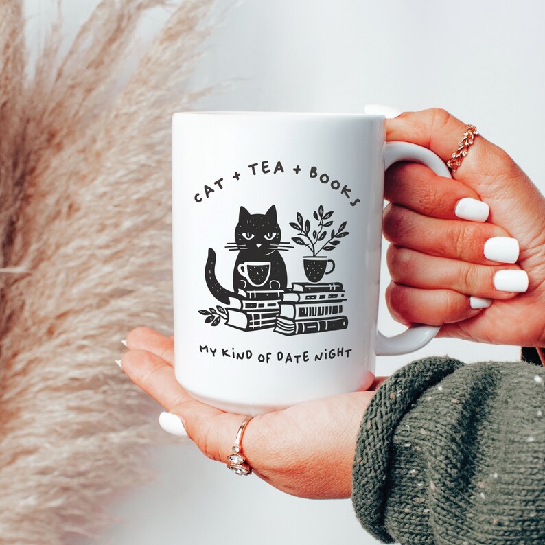 Puede incluir: Taza de cer&aacute;mica blanca con una ilustraci&oacute;n de un gato negro sentado en una pila de libros y una taza de t&eacute;. La taza tiene el texto "Cat + Tea + Books My Kind of Date Night" impreso en ella.
