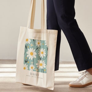 May include: A white canvas tote bag with a blue and white daisy print. The bag has the text "April Flower" and "Daisy" with the words "innocence, true love, cheerfulness" underneath.