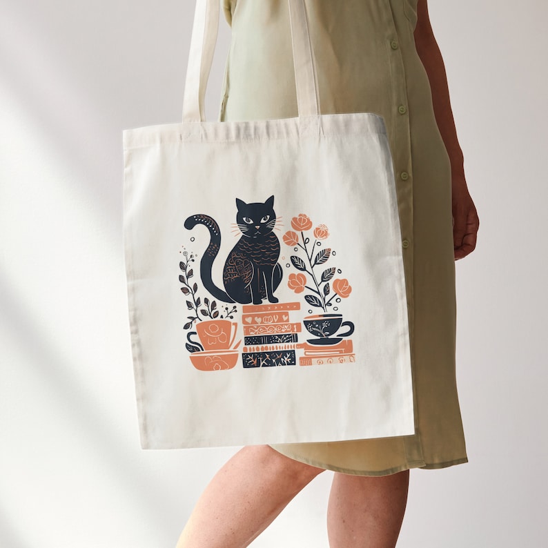 Puede incluir: Una bolsa de tela blanca con un gato negro sentado sobre una pila de libros y una taza. El gato est&aacute; rodeado de flores naranjas. El texto en la bolsa dice "So, so good."
