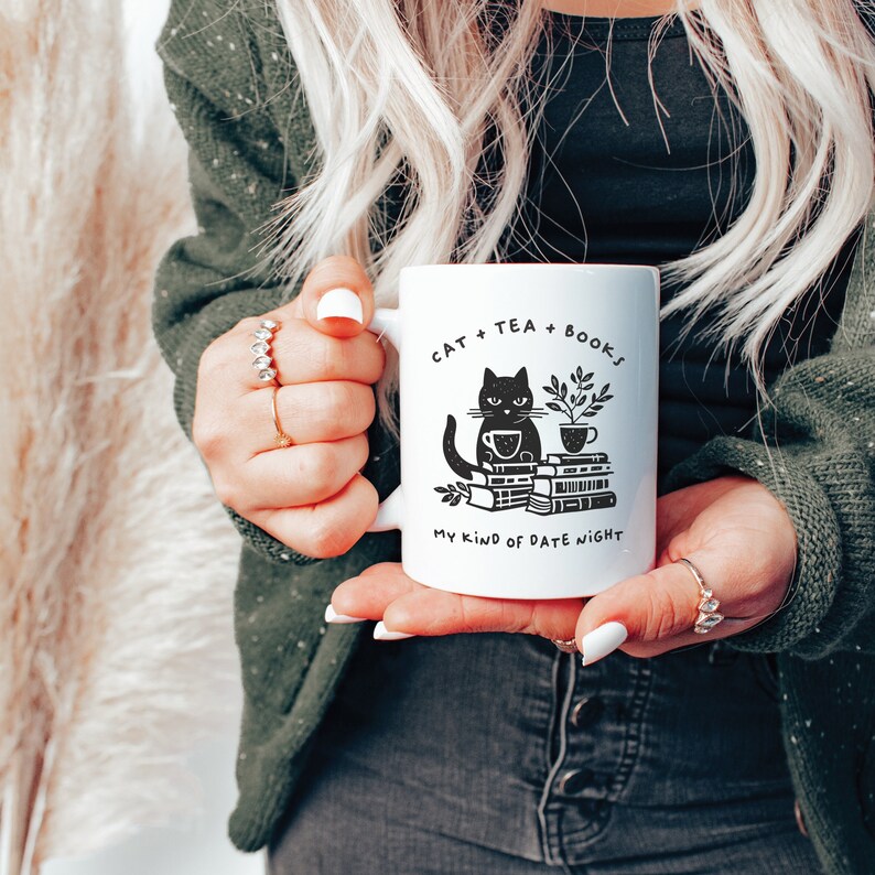 Puede incluir: Taza de cer&aacute;mica blanca con una ilustraci&oacute;n de un gato negro sosteniendo una taza de t&eacute; y sentado sobre una pila de libros. La taza tiene el texto "Cat + Tea + Books. My kind of date night."