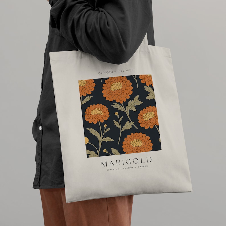 May include: A white tote bag with a black and orange marigold flower design. The bag has the text "October Flower" and "Marigold" with the words "Sympathy, Passion, Warmth" underneath.