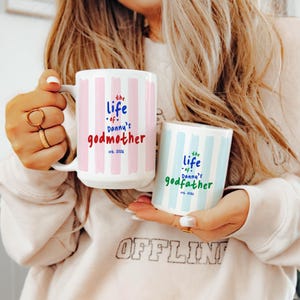 Könnte beinhalten: Zwei weiße Keramikbecher. Ein Becher hat rosa vertikale Streifen und den Text "the life of Danny's godmother est. 2026". Der andere Becher hat blaue Streifen und den Text "the life of Danny's godfather est. 2026".