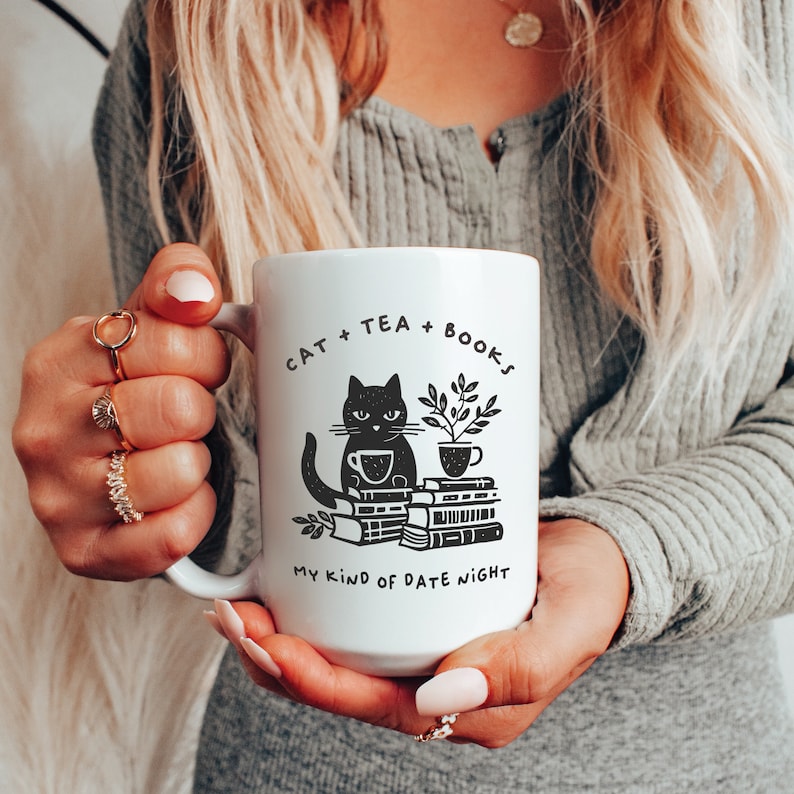 Puede incluir: Taza de cer&aacute;mica blanca con una ilustraci&oacute;n negra de un gato sentado en una pila de libros con una taza de t&eacute;. El texto en la taza dice "Cat + Tea + Books My Kind of Date Night".