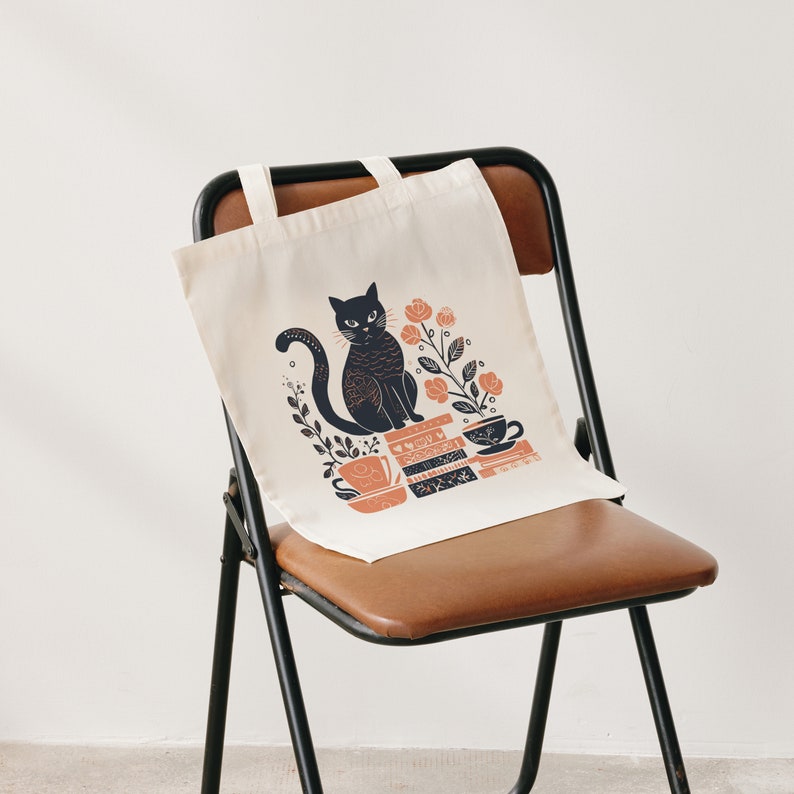 Puede incluir: Una bolsa de tela blanca con un gato negro sentado sobre una pila de libros. El gato est&aacute; rodeado de flores rosas y hojas verdes. La bolsa tiene un asa negra y est&aacute; sentada en una silla marr&oacute;n.