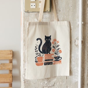 Puede incluir: Un gato negro sentado en una pila de libros con una taza de t&eacute; y flores. La bolsa de tela es de lona natural y presenta una ilustraci&oacute;n en blanco y negro.