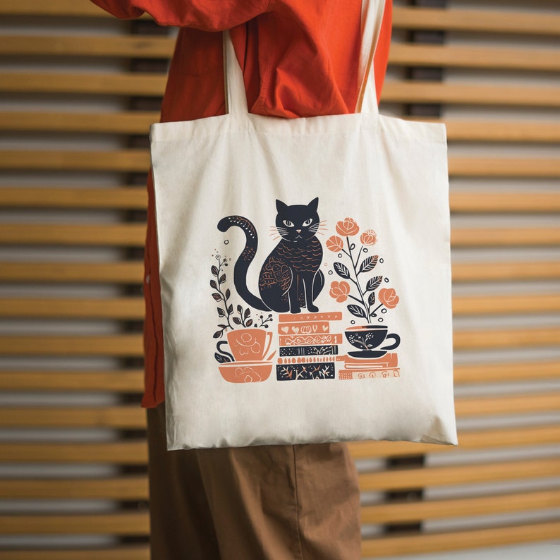 Puede incluir: Una bolsa de tela blanca con un gato negro sentado sobre una pila de libros. El gato est&aacute; rodeado de flores y una taza de t&eacute;. El texto en los libros dice "Love You More".