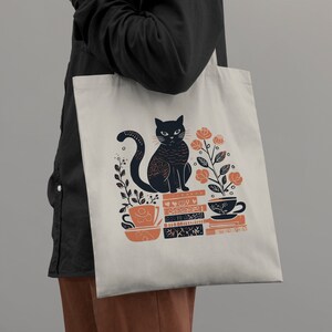 Puede incluir: Una bolsa de tela blanca con un gato negro sentado sobre una pila de libros. El gato est&aacute; rodeado de flores naranjas y hay dos tazas de t&eacute; en la pila de libros. El texto en la bolsa dice "Read, Sip, Repeat."