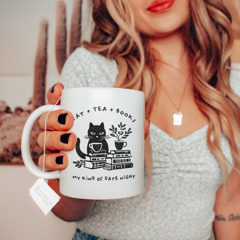 Puede incluir: Taza de cer&aacute;mica blanca con una ilustraci&oacute;n de un gato negro sentado sobre una pila de libros. La taza dice "Cat + Tea + Books My Kind of Date Night".