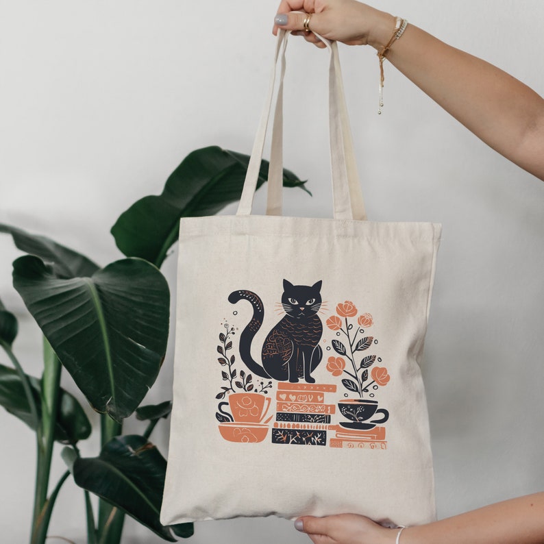 Puede incluir: Una bolsa de lona blanca con un gato negro sentado sobre una pila de libros y tazas de t&eacute;. El gato est&aacute; rodeado de flores y hojas. El texto en los libros es "The Book of Cats".