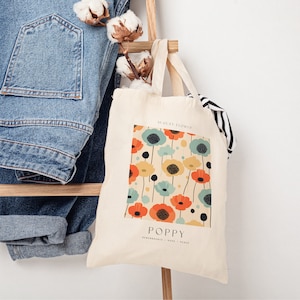 May include: A white canvas tote bag with a colorful poppy flower pattern. The bag has the text "August Flower" and "Poppy" printed on it. The text "Remembrance - Hope - Peace" is also printed on the bag.