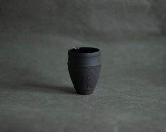 Puede incluir: Una pequeña taza de cerámica negra mate con una superficie exterior texturizada y rugosa y un acabado interior liso y brillante. El recipiente presenta una base cónica que se ensancha hacia el borde, creando una silueta muy distintiva.