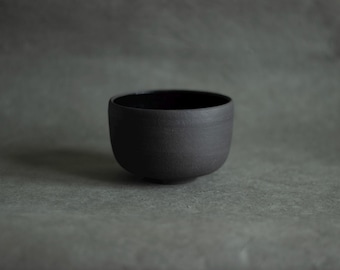 Puede incluir: Un cuenco de cerámica pequeño, redondo y de color negro mate, con un exterior liso y curvado y una parte superior abierta ligeramente más ancha. El interior del cuenco presenta un acabado oscuro y reflectante que contrasta con la superficie exterior texturizada.