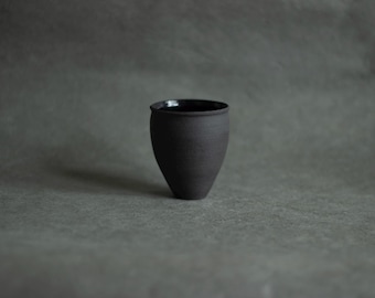 Puede incluir: Una pequeña taza de cerámica negra mate con una base cónica estrecha y un borde redondeado y liso. La superficie exterior presenta un acabado oscuro y texturizado, mientras que el interior parece tener un esmalte negro brillante.