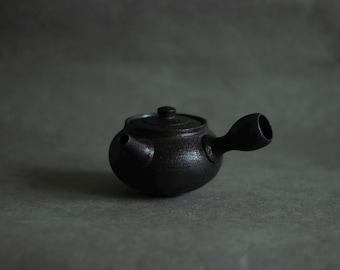 Puede incluir: Una tetera de color gris carbón oscuro con un acabado mate y texturizado. La tetera presenta un cuerpo redondeado, un pico pequeño, una tapa con un pomo central y un asa montada en el lateral que se extiende hacia afuera.