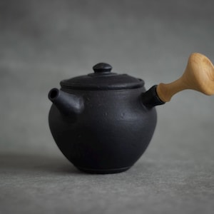 Könnte beinhalten: Eine mattschwarze Keramik-Teekanne mit Holzgriff und Deckel. Die Teekanne hat einen abgerundeten Körper und einen Ausguss an der Seite. Der Griff ist aus hellem Holz. Die Teekanne ist zum Aufbrühen von Tee bestimmt.