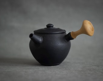 Puede incluir: Una tetera de cerámica negra mate con asa y tapa de madera. La tetera tiene un cuerpo redondeado y un pico en el lateral. El asa es de madera de color claro. La tetera está diseñada para preparar té.