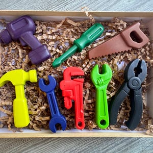 May include: A collection of colorful toy tools, including a purple drill, green screwdriver, brown saw, yellow hammer, blue wrench, red adjustable wrench, green wrench, and black pliers, arranged in a cardboard box with brown paper filler.
