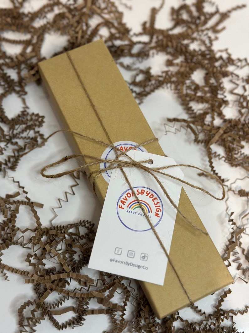 May include: A brown cardboard box tied with twine and a white tag. The tag has a logo that says "Favors By Design Party Favors" with a rainbow and the text "@FavorsByDesignCo".