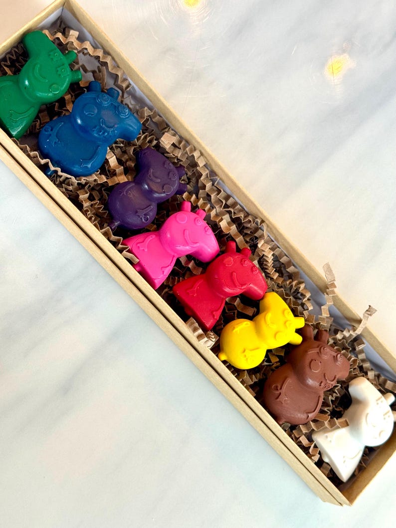 May include: A set of eight colorful crayons shaped like Peppa pig, packaged in a brown box with shredded paper. The crayons are arranged in a rainbow order, starting with green and ending with white.
