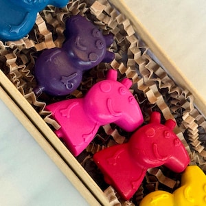 May include: A set of five colorful crayons shaped like cartoon pigs in a brown cardboard box with brown crinkle paper. The crayons are blue, purple, pink, red, and yellow.
