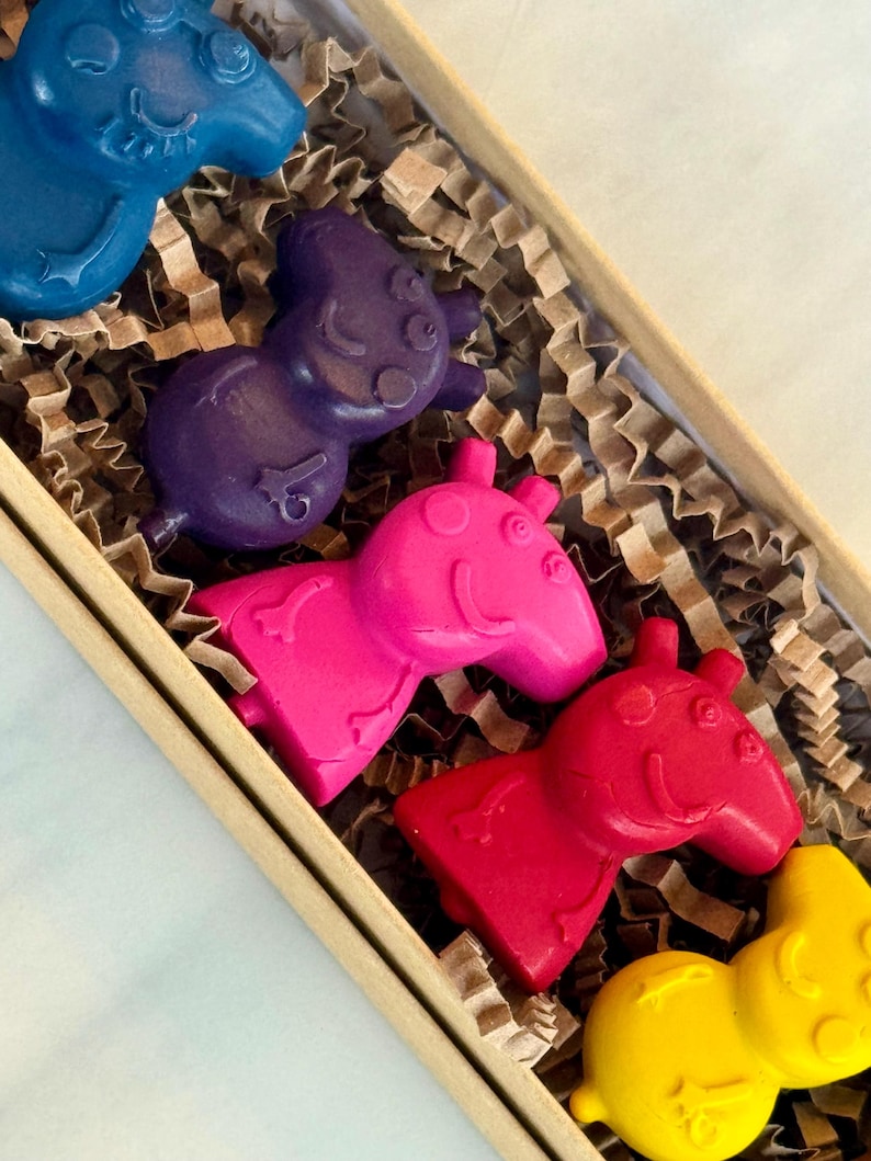 May include: A set of five colorful crayons shaped like cartoon pigs in a brown cardboard box with brown crinkle paper. The crayons are blue, purple, pink, red, and yellow.