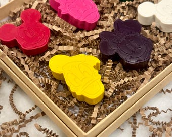 May include: A set of character-shaped crayons in a square box filled with brown paper shreds. The crayons are in various colors, including red, pink, green, white, yellow, and purple. The crayon shapes resemble cartoon characters.