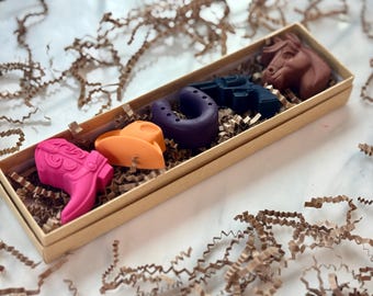 May include: A decorative box filled with colorful cowboy-themed wax figures. The box contains a pink cowboy boot, an orange cowboy hat, a purple horseshoe, a blue revolver, and a brown horse head. The box is surrounded by brown crinkle paper.