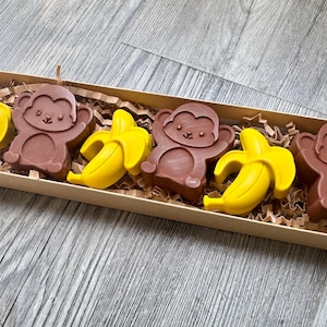 May include: A rectangular box contains six chocolate treats. Three brown monkey-shaped chocolates alternate with three yellow banana-shaped chocolates. The box is filled with brown crinkle paper.