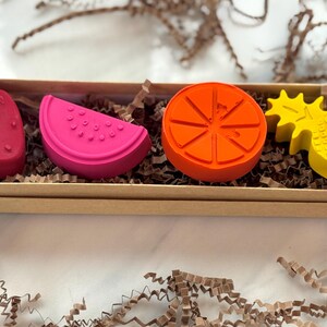 May include: A set of four fruit-shaped crayons in a rectangular box. The crayons are a strawberry, watermelon slice, orange slice, and pineapple. The crayons are red, pink, orange, and yellow. The box is filled with brown crinkle paper.